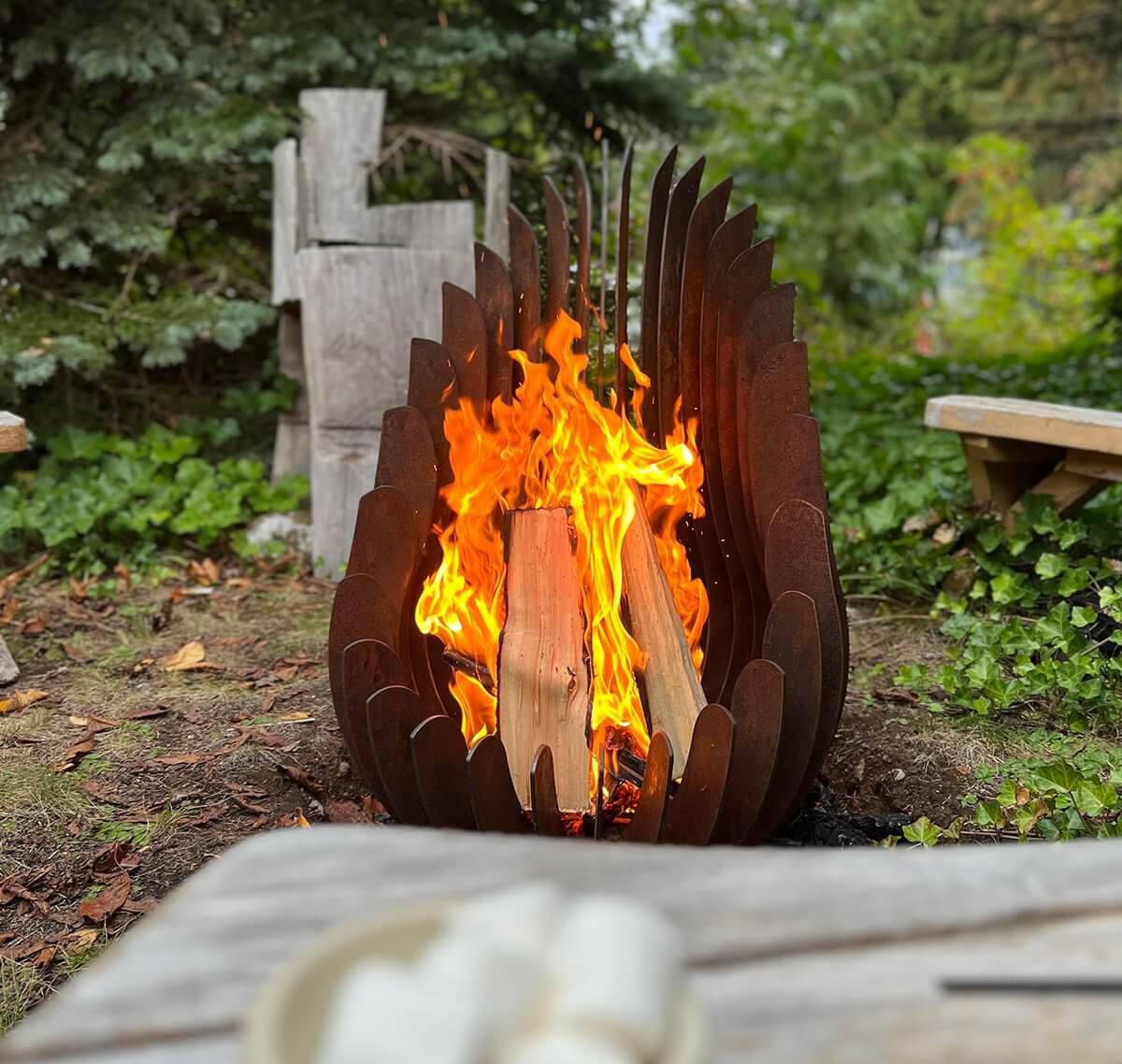 Kelray sculptural firepit burning wood in a forested backyard setting, with marshmallows and rustic seating nearby